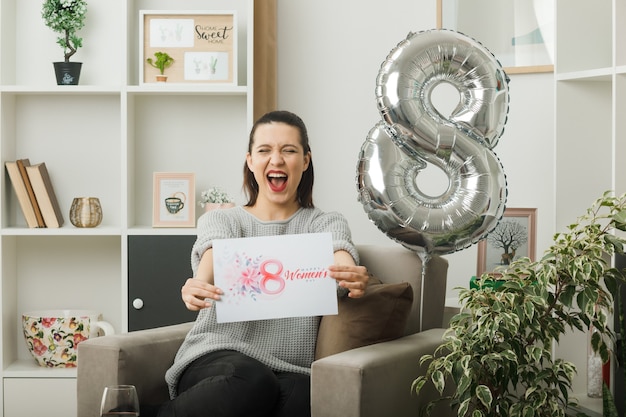 Opgewonden mooi meisje op gelukkige vrouwendag stak een wenskaart uit, zittend op een fauteuil in de woonkamer