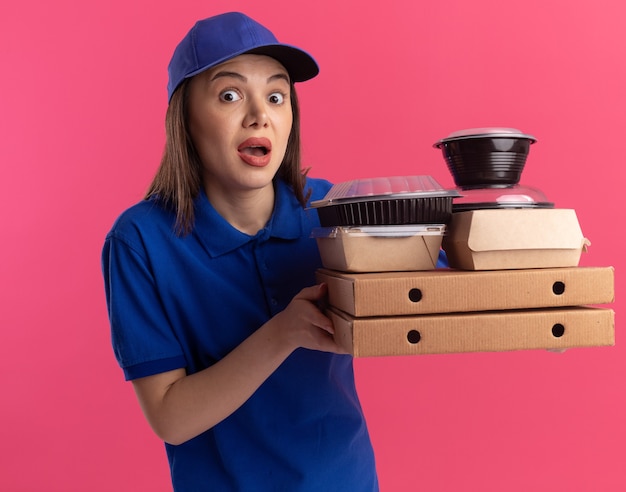 Opgewonden mooie bezorger in uniform houdt voedselpakket en containers op pizzadozen en geïsoleerd op roze muur met kopieerruimte