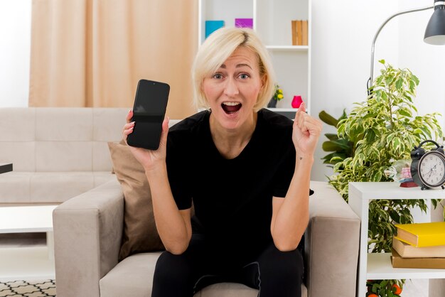 Opgewonden mooie blonde Russische vrouw zit op fauteuil vuist houden en telefoon in de woonkamer te houden