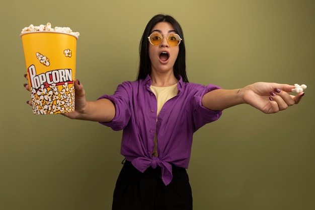Opgewonden mooie brunette vrouw in zonnebril houdt emmer popcorn geïsoleerd op olijfgroene muur