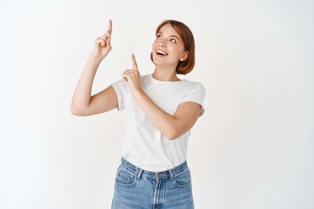 Opgewonden mooie dame in t-shirt die met de vingers omhoog wijst, glimlacht en er geamuseerd uitziet, promotieadvertentie bekijkt, op een witte muur staat
