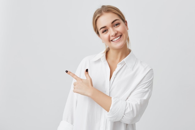 Opgewonden mooie jonge blonde blanke vrouw draagt een wit overhemd met haar wijsvinger zijwaarts wijzend, vrolijk lachend met haar witte tanden, en toont iets verrassends op een grijze kopie ruimte muur