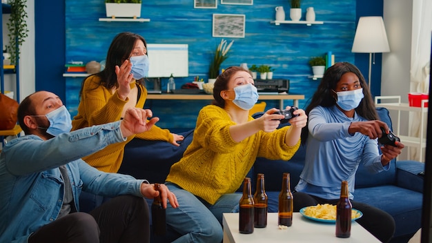 Opgewonden multi-etnische vrienden die videogames proberen te winnen en genieten van een nieuw normaal feest tijdens de wereldwijde pandemie met gezichtsmasker, afstand houdend zittend op de bank in de woonkamer en vrouwen ondersteunend