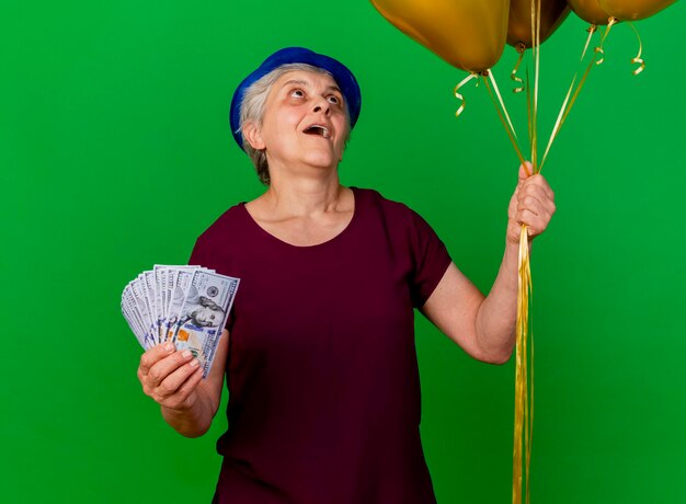 Opgewonden oudere vrouw met feestmuts houdt geld vast en kijkt naar helium ballonnen op groen