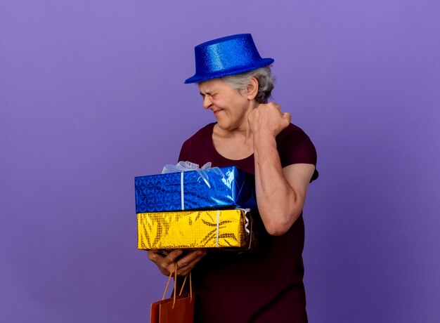 Opgewonden oudere vrouw met feestmuts houdt geschenkdozen en houdt vuist geïsoleerd op paarse muur met kopie ruimte