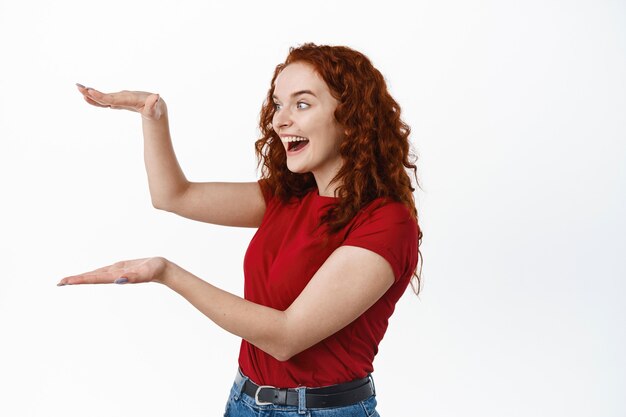 Opgewonden roodharige meid die een groot object laat zien, lege ruimte met handen vasthoudt, verbaasd naar het product staart, tegen een witte muur staat