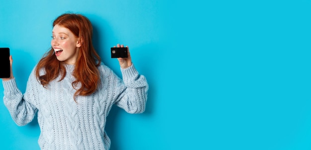 Gratis foto opgewonden roodharige meid die het scherm van de mobiele telefoon laat zien en een creditcard die een online winkel of app aantoont