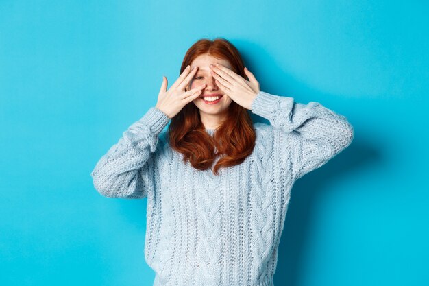 Opgewonden roodharige tienermeisje opent ogen om vakantieverrassing te zien, cadeautjes te ontvangen, verbaasd naar de camera te kijken, staande over blauwe achtergrond.