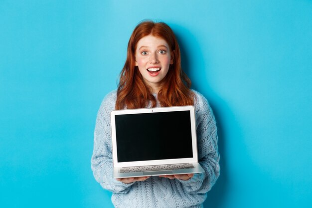 Opgewonden roodharige vrouwelijke freelancer die een laptopscherm toont, verbaasd naar de camera staart, met de computer tegen een blauwe achtergrond staat