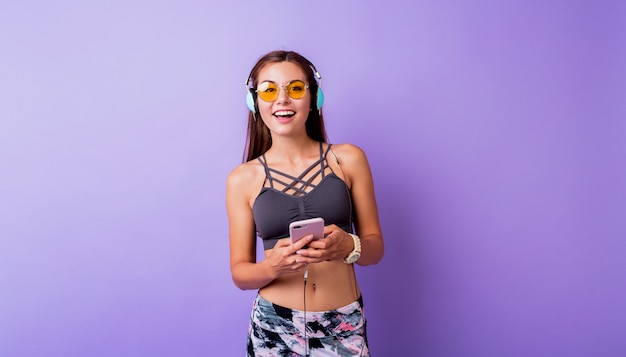 Opgewonden sportieve vrouw met oprecht glimlachend poseren in stijlvolle actieve slijtage op studio. smartphone gebruiken en muziek luisteren.