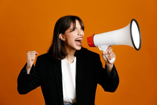 opgewonden spreekt op luidspreker jonge mooie vrouw draagt zwarte jas geïsoleerd op oranje achtergrond