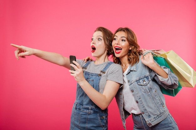 Opgewonden twee vrouwenvrienden die boodschappentassen houden gebruikend mobiele telefoon.