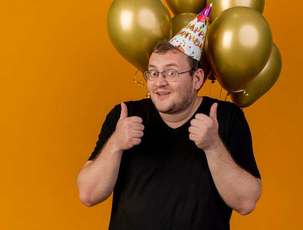 Opgewonden volwassen slavische man in optische bril met verjaardagspet staat voor heliumballonnen en duimen omhoog met twee handen