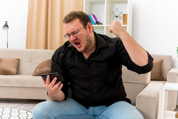 Opgewonden volwassen Slavische man in optische bril zit op fauteuil met opgeheven vuist omhoog kijken naar telefoon in de woonkamer