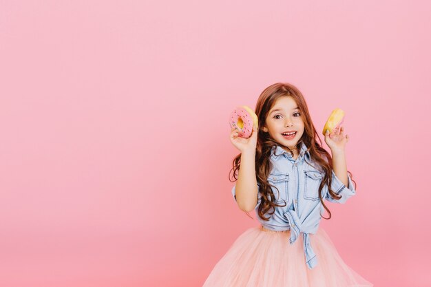 Opgewonden vrolijk jong mooi meisje in tule rok positiviteit uiten, plezier voor camera met donuts geïsoleerd op roze achtergrond. Gelukkige jeugd met lekker dessert. Plaats fot tekst