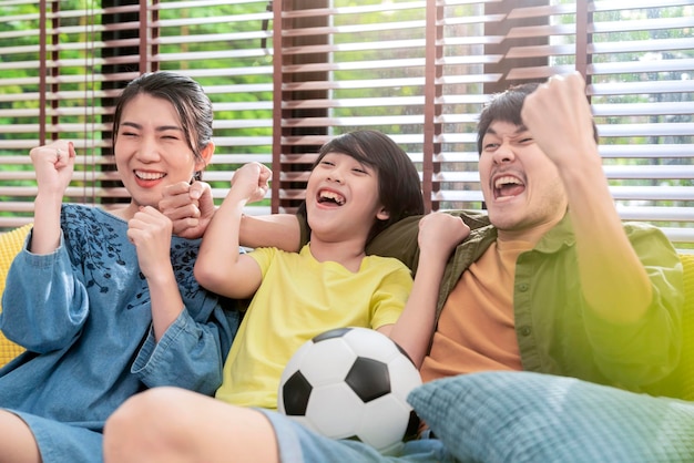 Opgewonden vrolijke blije Aziatische ouders en zoon kijken naar sport op tv juichen favoriete voetbalteam samen zitten op de bankbank thuis Weekend met vader Familie tijd Online tv-concept