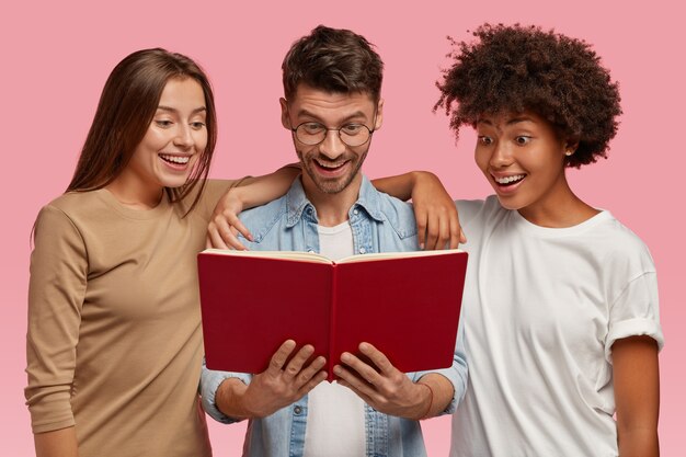 Opgewonden vrolijke nieuwsgierige multi-etnische twee jonge vrouwen en knappe jongen kijken naar leerboek, lezen informatie, geïsoleerd over roze muur. Gelukkige interraciale studenten leren iets voor het examen