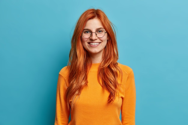 Opgewonden vrolijke roodharige vrouw met een oprechte brede glimlach heeft plezier en ziet er direct uit met een ronde optische bril gekleed in een oranje trui.