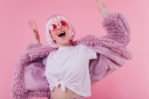 Opgewonden wit vrouwelijk model in bontjas dansen in studio Indoor foto van emotioneel meisje in zonnebril en roze periwig lachen tijdens fotoshoot