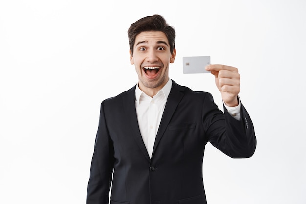 Opgewonden zakenman toont creditcard en glimlacht, geopende storting, staande tegen witte muur in zwart pak