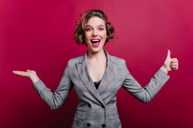 Opgewonden zakenvrouw in elegante grijze kleding met plezier tijdens fotoshoot. enthousiast meisje in formele stijl jas zwaaiende handen op bordeaux muur.