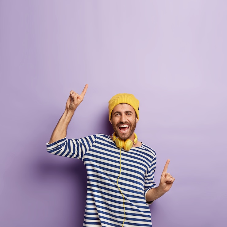 Gratis Foto opgewonden zorgeloze positieve man in casual gestreepte trui, gele hoed, wijst vrolijk omhoog op geweldige kopie ruimte voor je promo