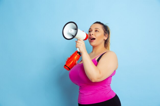 Opleiding van het jonge Kaukasische plus grootte vrouwelijke model op blauwe achtergrond. Concept van sport, menselijke emoties, expressie, gezonde levensstijl, positief lichaam, gelijkheid. Poseren met fles en mondvrede.