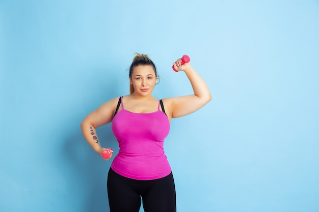 Opleiding van het jonge Kaukasische plus grootte vrouwelijke model op blauwe achtergrond. Concept van sport, menselijke emoties, expressie, gezonde levensstijl, positief lichaam, gelijkheid. Trainen met de gewichten, copyspace.