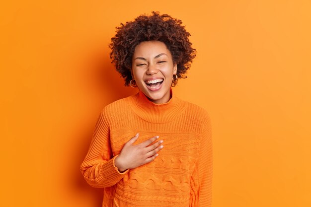 Oprechte emoties en positieve gevoelens concept. Gelukkig tevreden vrouw glimlacht houdt in het algemeen hand op de borst lacht om grappig verhaal gekleed in casual trui geïsoleerd over oranje muur