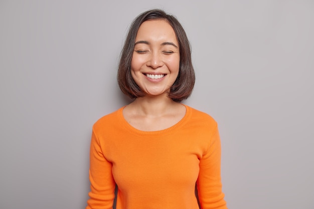 Oprechte mooie Aziatische vrouw blij om hartverwarmende woorden te horen houdt ogen gesloten glimlacht zachtjes heeft natuurlijk donker haar gezonde huid gekleed in casual oranje trui poses tegen grijze muur