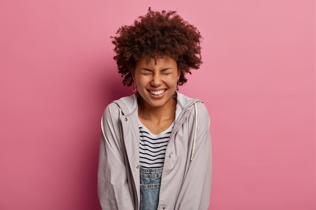 Gratis foto oprechte, vrolijke vrouw met krullend haar heeft een brede stralende glimlach, geniet van het leven zonder problemen, lacht om grappige grapjes, loenst gezicht met plezier, draagt een casual anorak, poseert over een roze muur.