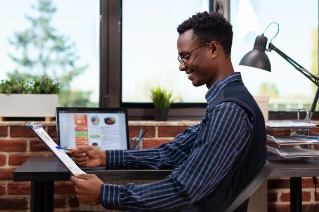 Opstarteigenaar met klembord met jaarlijkse bedrijfsgrafieken blij met omzetresultaten aan bureau met laptop. Ondernemer die key performance indicators op draagbare computer vergelijkt.