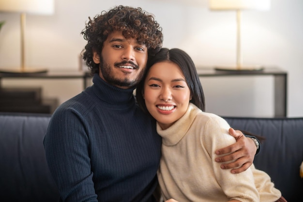 Optimisten. Aantrekkelijke jonge, bebaarde Indiase man die een schattige aziatische vrouw omhelst met lang donker haar en vol vertrouwen naar de camera kijkt die thuis op de bank zit