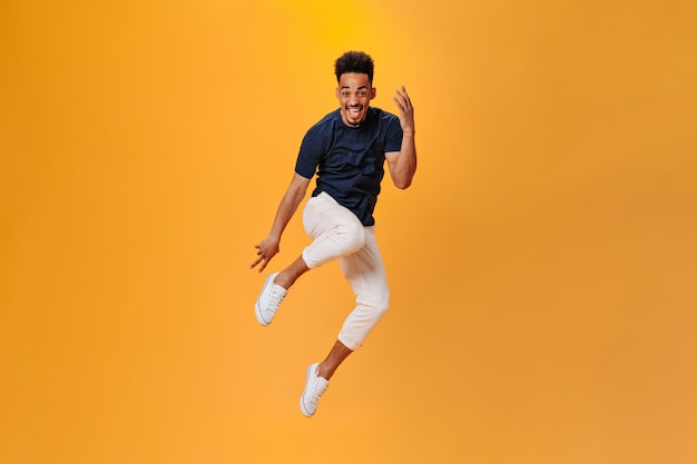 Optimistische donkere gevilde man in stijlvolle outfit springen op oranje achtergrond Brunette man in zwarte t-shirt en witte broek poseren op geïsoleerde