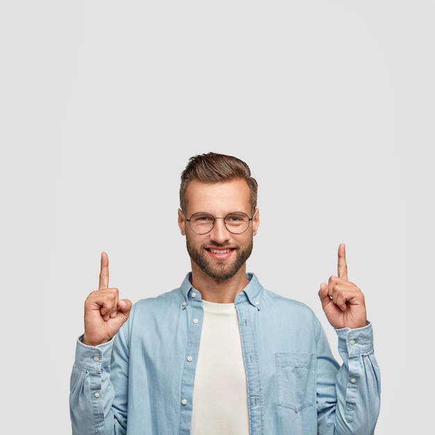 Optimistische ongeschoren jongeman heeft trendy kapsel, opgewekte uitdrukking, wijst met beide wijsvingers naar boven en is in een goed humeur