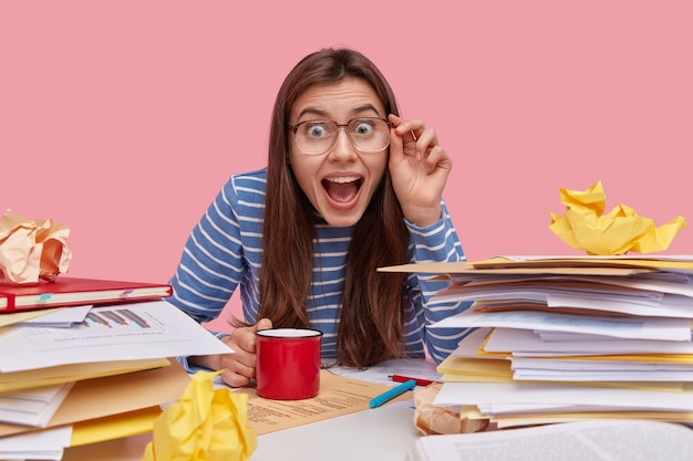 Optimistische vrolijke dame met donker haar, houdt de hand op de brilrand, geopende mond, gekleed in gestreepte kleding, drinkt warme dranken, werkt in de coworking-ruimte