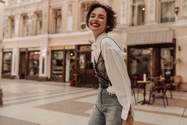 Optimistische vrouw met krullend haar in spijkerbroek oprecht glimlachen in de stad. Stoere dame in lichte blouse met zwart kant op straat.