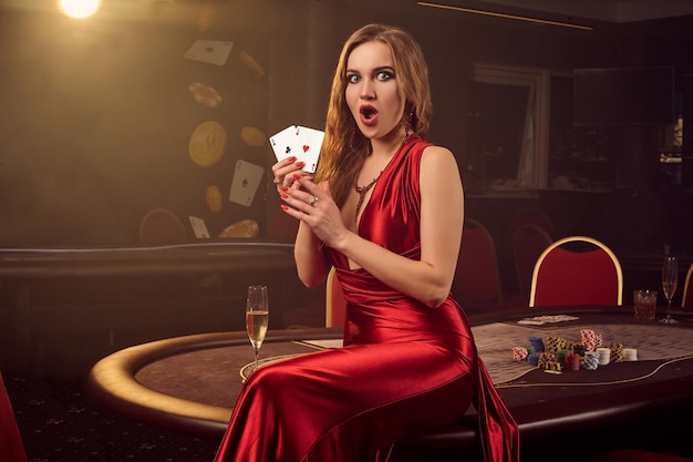 Opzichtige blonde vrouw in een lange rode satijnen jurk, met twee azen in haar handen kijkt verbaasd en poseert zittend op een pokertafel in een luxe casino. Passie, kaarten, chips, alcohol, winnen, gokken - het is
