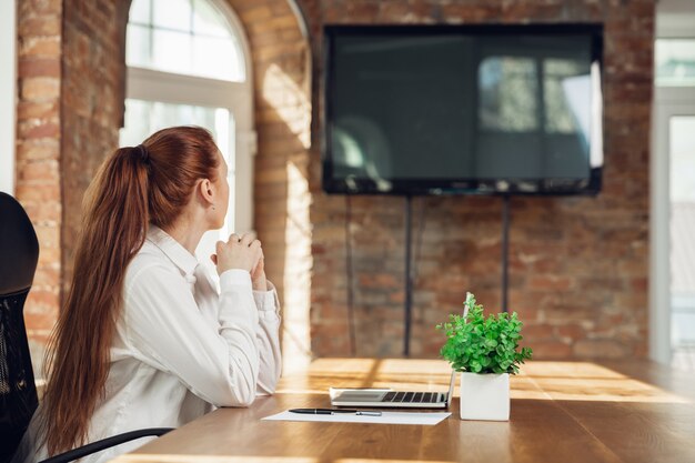 Opzij kijken. Blanke jonge vrouw in zakelijke kleding die op kantoor werkt. Jonge zakenvrouw, manager die taken doet met smartphone, laptop, tablet heeft online conferentie. Concept van financiën, baan.