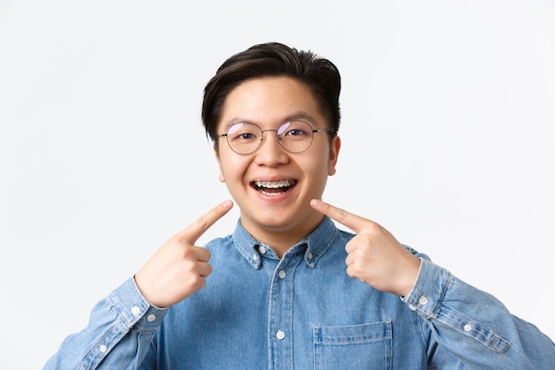 Orthodontie en stomatologie concept. Close-up van tevreden aziatische man, klant van de tandheelkundige kliniek die blij glimlacht en naar zijn beugel wijst, op een witte achtergrond staat, kwaliteit aanbeveelt.