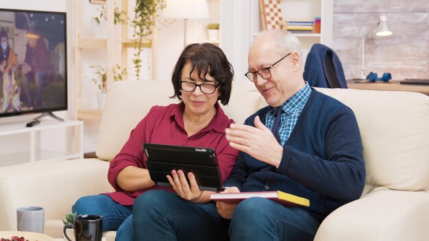 Oud koppel zittend op de bank tijdens een videogesprek op tablet. Oud paar dat bij tablet zwaait.