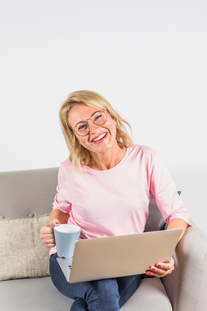 Oude lachende vrouw in roze blouse met laptop en kop op bank