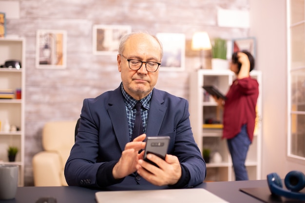 Oude man die nieuwe technologie gebruikt in een gezellig huis. Hij heeft een smartphone in zijn handen