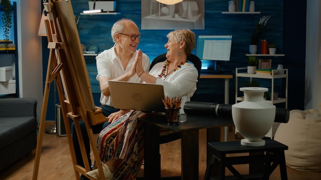 Oude man en vrouw die samenwerken om vaaskunstwerken te tekenen, met behulp van een laptopcomputer om online moderne inspiratie te vinden. team van kunstenaars tekenen met artistieke hulpmiddelen en creatieve vaardigheden, schets meesterwerk.