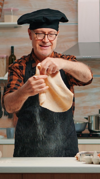 Oude man met keukenschort spelen met brooddeeg thuis glimlachend voor camera. Gepensioneerde oudere chef-kok die pizza-aanrecht vormt op een met bloem bestoven oppervlak en het met de handen kneedt, in moderne keuken