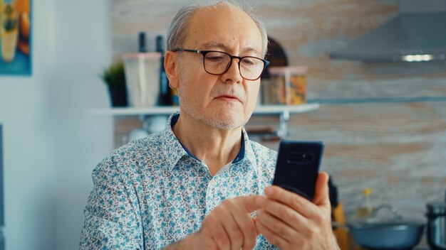 Oude man surfen op sociale media met smartphone tijdens het ontbijt in de keuken. Authentiek portret van gepensioneerde senior die geniet van moderne online internettechnologie