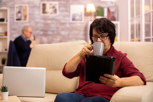 Oude oudere vrouw die op de bank zit en een digitale tablet-pc gebruikt in een gezellige woonkamer.