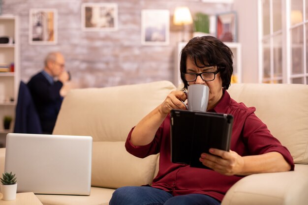 Oude oudere vrouw die op de bank zit en een digitale tablet-pc gebruikt in een gezellige woonkamer.