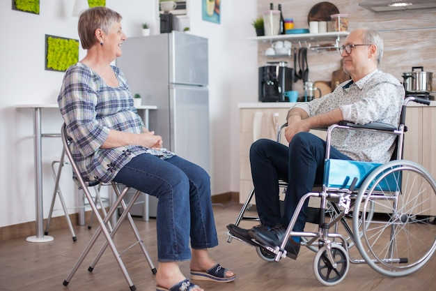 Oude vrouw en haar gehandicapte man in rolstoel chatten in de keuken. Bejaarde die een gesprek heeft met de man in de keuken. Leven met een gehandicapte met een loophandicap