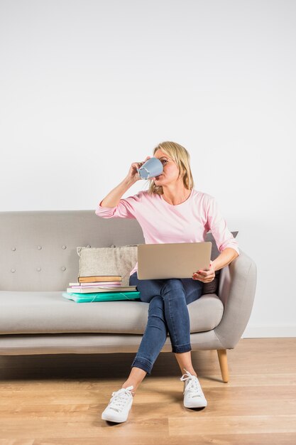 Oude vrouw in roze blouse met laptop en hoop van boeken die van kop op bank drinken